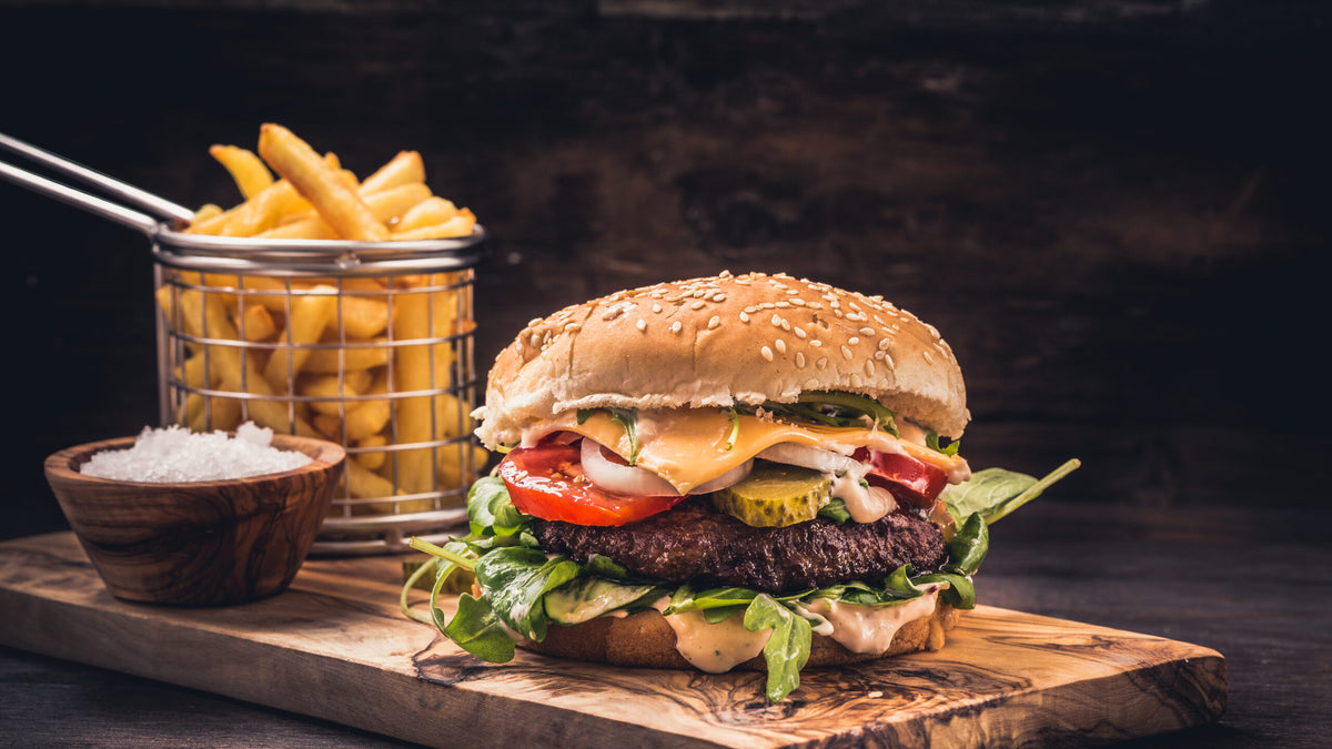 Saftiger, appetitlich angerichteter Gourmet-Burger mit frischem Salat, Tomaten, Käse, Gurken und cremiger Sauce, serviert auf einem rustikalen Holzbrett. Im Hintergrund steht ein Metallkorb mit knusprigen Pommes frites, angeboten im amerikanischen All-You-Can-Eat-Buffet von ISM Foodspot Dortmund.
