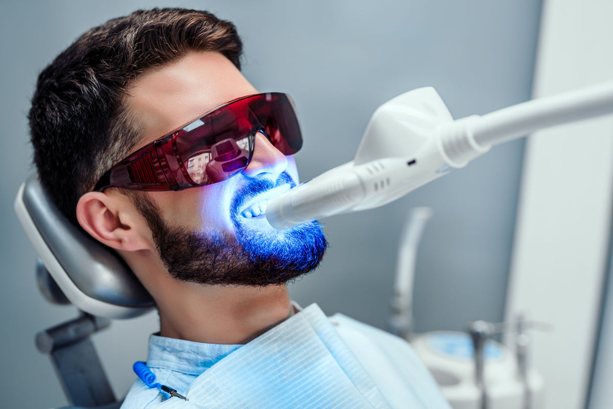 Ein Mann mit Schutzbrille erhält eine professionelle Zahnbleaching-Behandlung. Seine Zähne werden mit einem speziellen Blaulicht bestrahlt, um die Aufhellung der Zähne zu unterstützen.