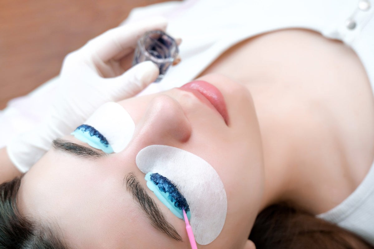 Nahaufnahme einer liegenden Frau während einer professionellen Wimpernfärbung. Ihre Augen sind geschlossen und mit Schutzpads versehen, während eine Kosmetikerin mit Handschuhen die Farbe sorgfältig auf die Wimpern aufträgt.