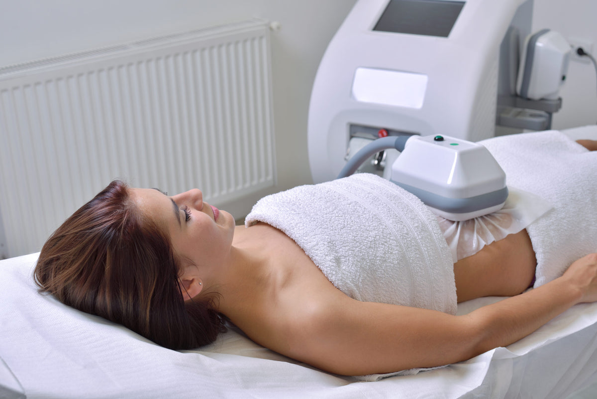 Junge Frau liegt entspannt auf einer Liege, während sie eine Cryolipolyse-Behandlung am Bauchbereich erhält. Das Behandlungsgerät ist auf dem Bauch fixiert.