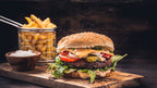Saftiger Cheeseburger mit Fleischpatty, Tomaten, Gurken, Zwiebeln, Salat und Sauce im Sesambrötchen, serviert mit Pommes in Metallkörbchen auf Holzbrett