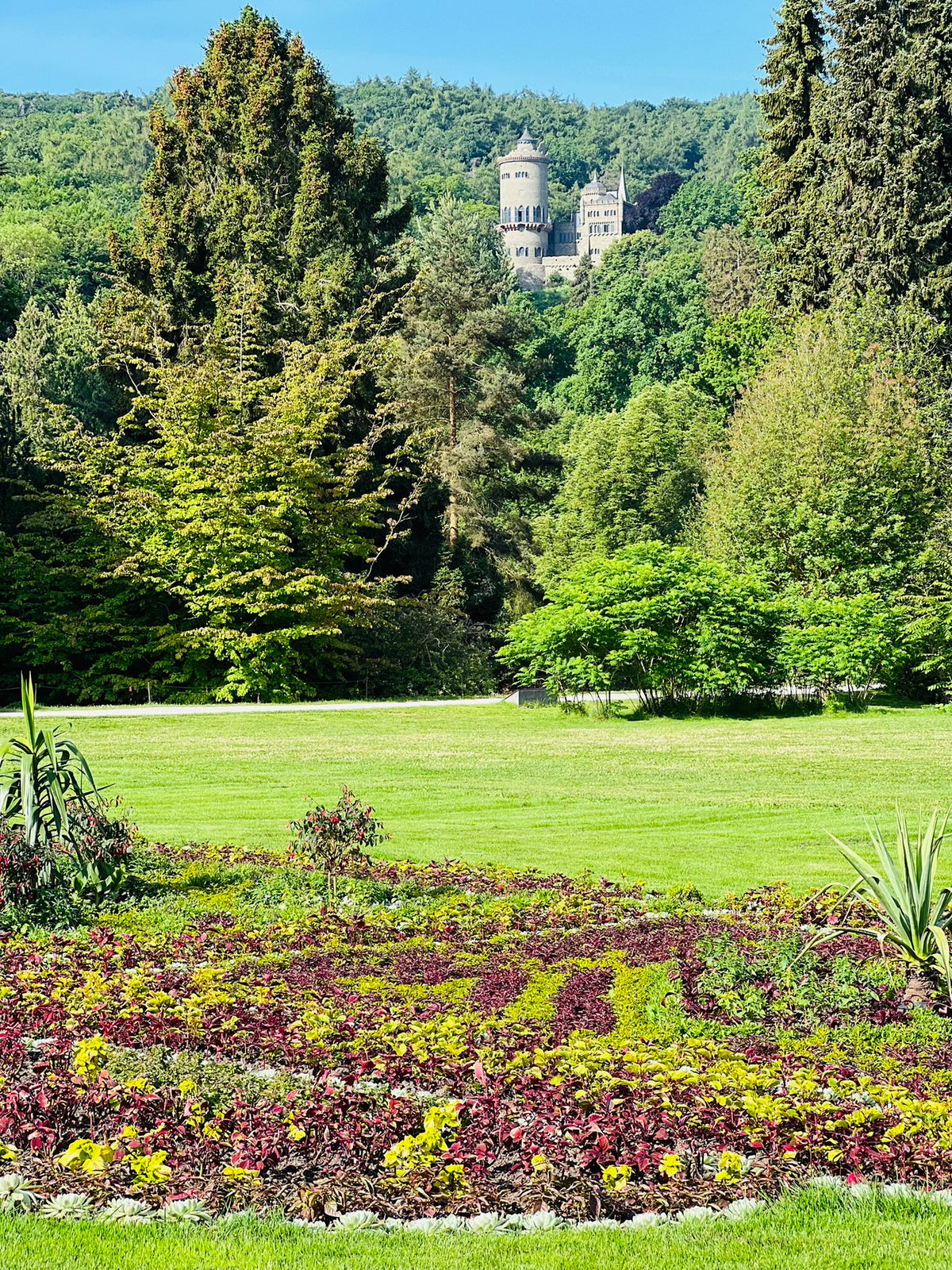 Grüne Gartenanlage im Bergpark Wilhelmshöhe mit blühenden Beeten im Vordergrund, dichtem Baumbestand und der Herkulesstatue in der Ferne