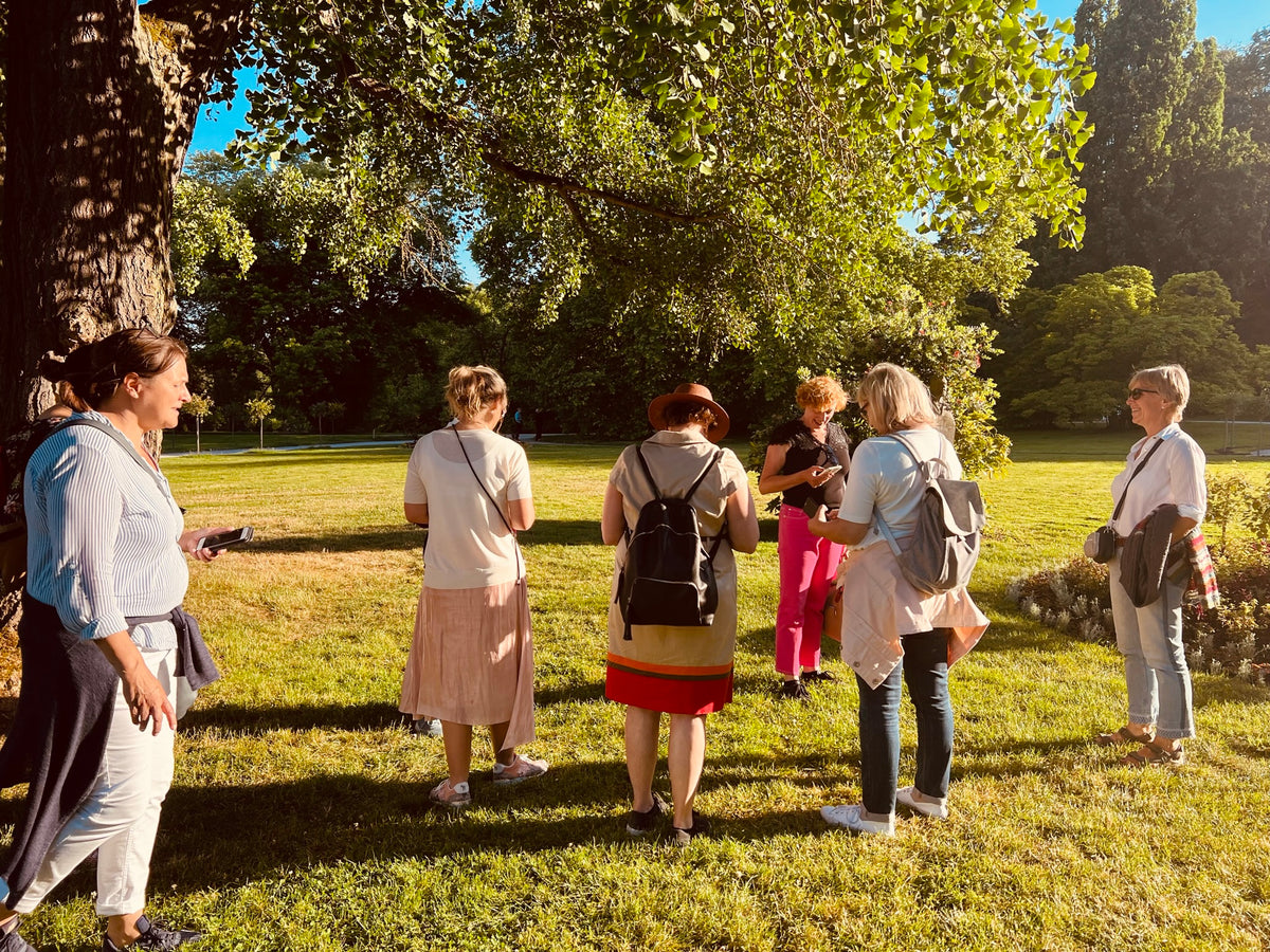 Gruppe von Stadtkrimi-Teilnehmenden im Park bei sonnigem Wetter, diskutieren über Hinweise mit Smartphone und Roadbook in der Hand unter Bäumen