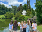 Jugendgruppe der Stadtkrimis Kassel steht mit Rätselheft am Seeufer im Bergpark und blickt auf den weißen Tempel-Pavillon, eingebettet in grüne Landschaft