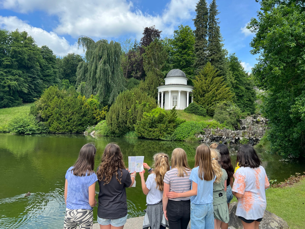 Jugendgruppe der Stadtkrimis Kassel steht mit Rätselheft am Seeufer im Bergpark und blickt auf den weißen Tempel-Pavillon, eingebettet in grüne Landschaft