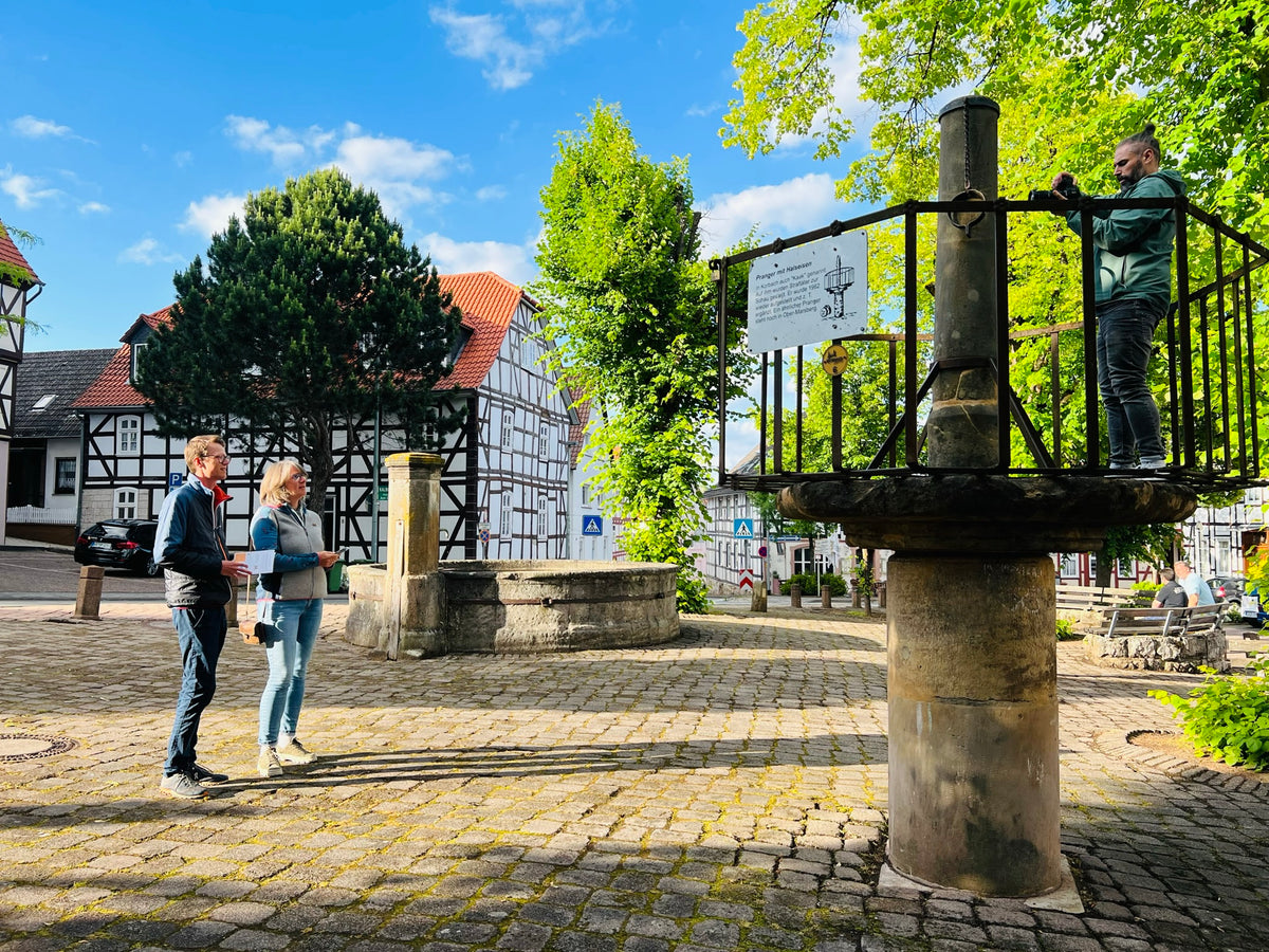 Zwei Stadtkrimi-Teilnehmende stehen an einem historischen Brunnenplatz mi Fachwerkhäusern im Hintergrund, bei sonnigem Wetter in Kassel auf Spurensuche