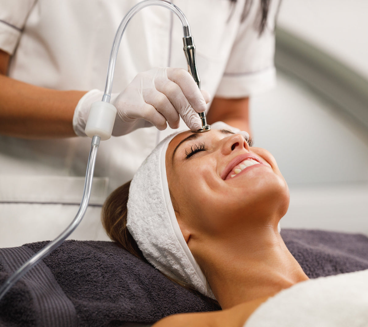 Frau liegt entspannt auf der Behandlungsliege und erhält eine Microdermabrasion im Stirnbereich mit speziellem Gerät durch eine Kosmetikerin in Weiß