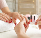 Nageldesignerin trägt roten Lack auf frisch modellierte Fingernägel einer Kundin auf, im Hintergrund stehen weitere Nagellacke auf dem Tisch