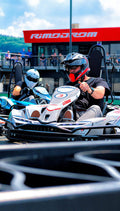 Nahaufnahme eines Fahrers mit Helm im Kart auf der Strecke des Rimodrom Hemer, ein zweiter Fahrer im Hintergrund, Zuschauerbereich und Tribüne leicht unscharf sichtbar