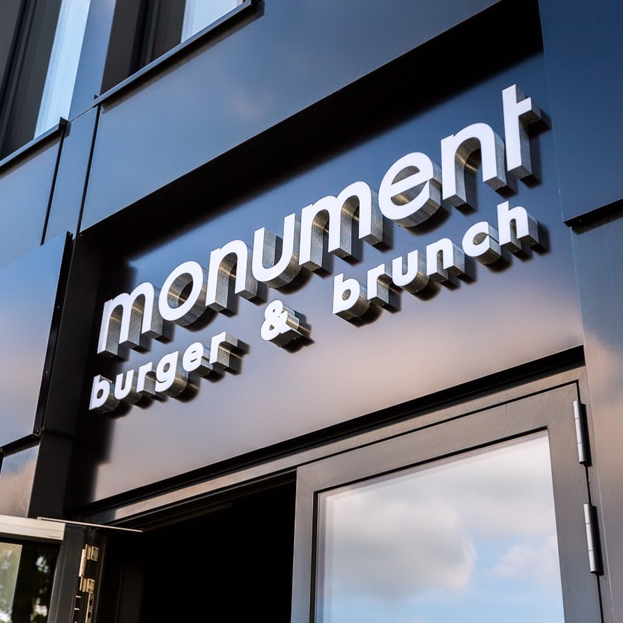 Eingangsbereich von Monument – Burger & Brunch mit modernem Fassaden-Schriftzug in silbernen Lettern auf dunklem Hintergrund