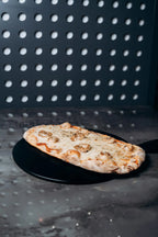 Pinsa mit geschmolzenem Käse und Garnelen, serviert auf schwarzem Teller vor moderner gepunkteter Wand im Shiva Food and More Dinslaken