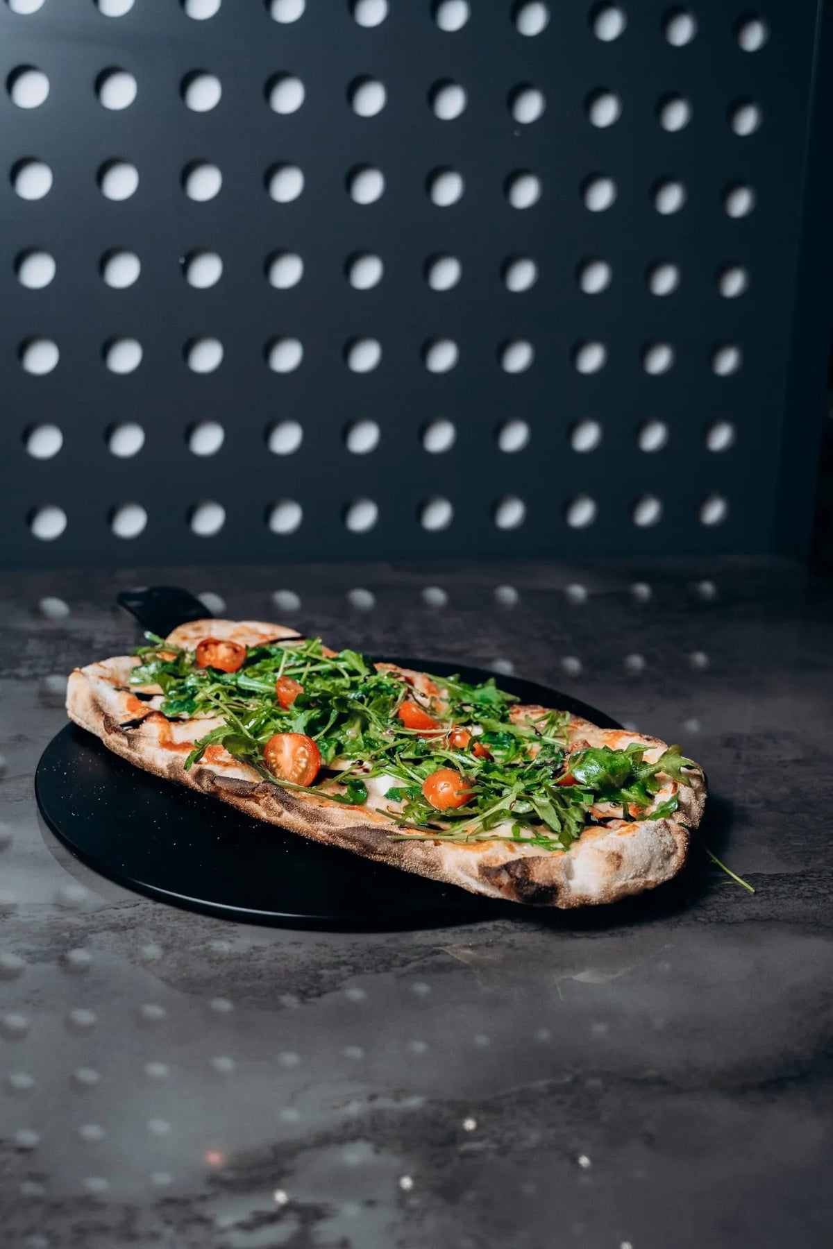 Ofenfrische Pinsa vom Shiva Food and More Dinslaken, belegt mit Rucola, Cherrytomaten und cremiger Basis, serviert auf dunklem Teller vor moderner Wandkulisse