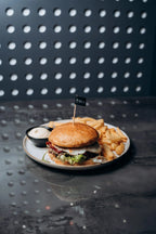 Burger mit Sesam-Bun, Salat, Bacon, Käse und Sauce, serviert mit Pommes frites und Dip auf einem Teller im Shiva Food and More Dinslaken, vor moderner gepunkteter Wand