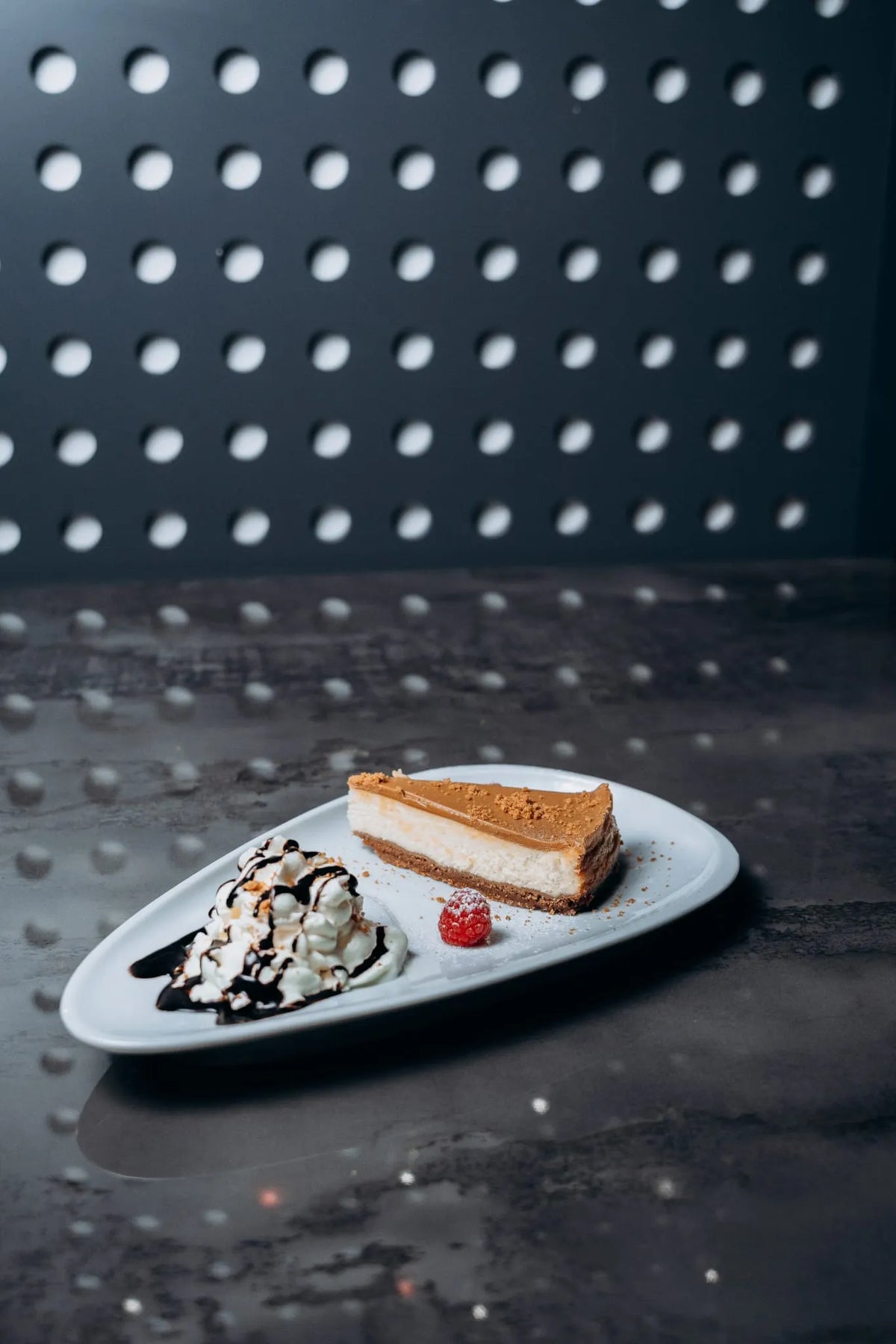 Dessertteller mit Cheesecake, einer Kugel Vanilleeis mit Schokosauce, Sahne und Himbeere, stilvoll angerichtet auf moderner Platte im Shiva Food and More Dinslaken