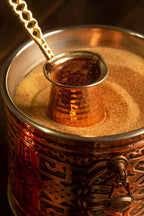 Kupferkännchen mit Schwarztee im heißen Sandbad, aromatisiert mit Kardamom – traditionelle Zubereitung in einem verzierten Gefäß