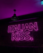 Leuchtendes pinkes Neon-Logo „ITALIAN KISS.“ an einer Backsteinwand im Innenraum des Italian Kiss Köln, inszeniert mit stimmungsvoller Beleuchtung