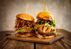 Zwei saftige BBQ-Burger auf einem Holzbrett aus dem Red Barn BBQ Restaurant, gefüllt mit Fleisch, Salat, Käse, Zwiebelringen und BBQ-Sauce