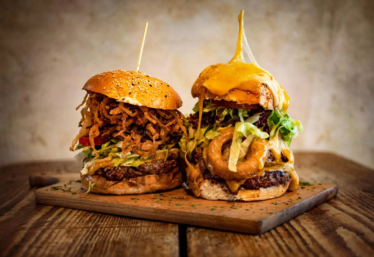 Zwei saftige BBQ-Burger auf einem Holzbrett aus dem Red Barn BBQ Restaurant, gefüllt mit Fleisch, Salat, Käse, Zwiebelringen und BBQ-Sauce