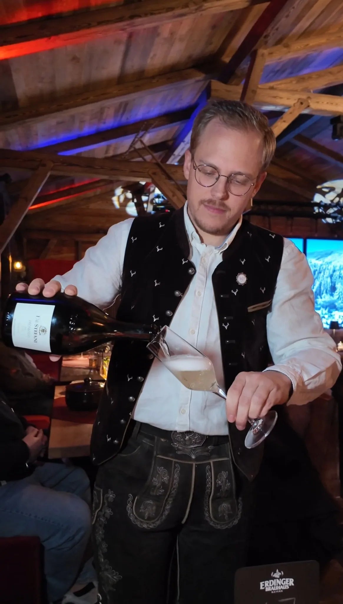 Servicemitarbeiter in traditioneller Tracht schenkt stilvoll Wein ein bei Abendveranstaltung in der Alm Fernweh, im Hintergrund rustikale Almhütten-Atmosphäre