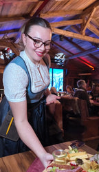 Lächelnde Servicemitarbeiterin in Dirndl serviert Speisen bei Abendveranstaltung in der Alm Fernweh, im Hintergrund rustikales Ambiente mit Holzbalken und Gästen