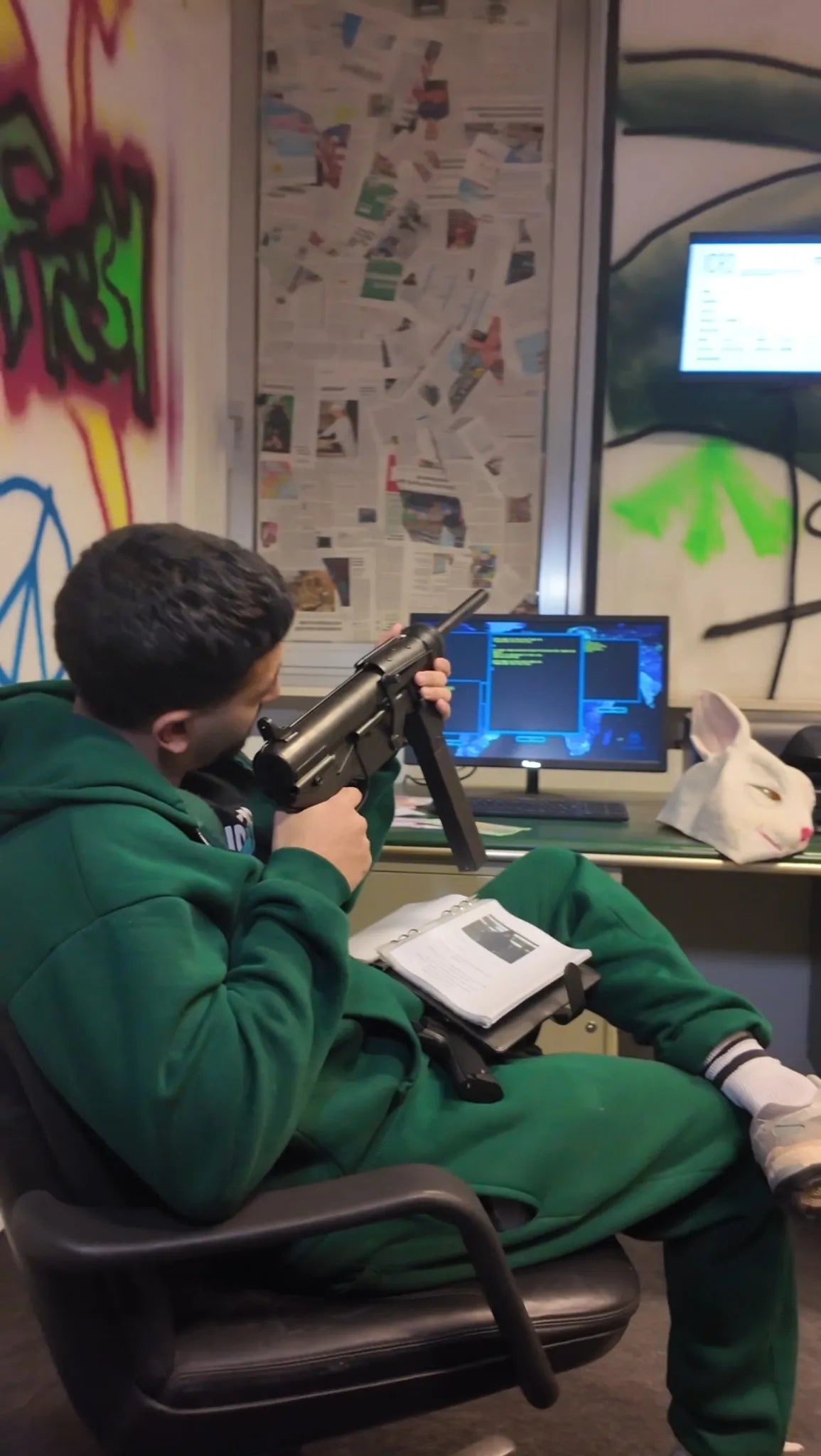 Teilnehmer in grüner Kleidung sitzt im Hacker-Raum bei Face Off Rüsselsheim mit Spielzeugwaffe, Buch, Hasenmaske und Bildschirm voller Codedaten im Hintergrund