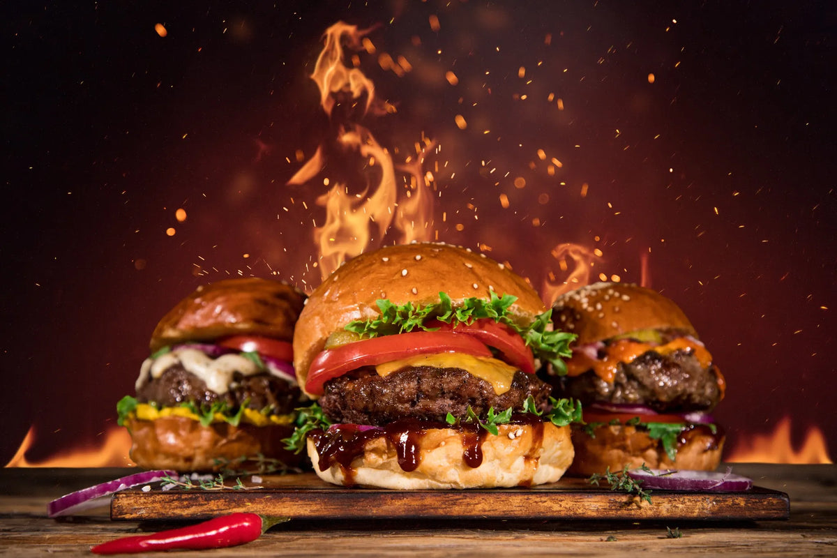 Drei saftige Burger mit verschiedenen Toppings wie Käse, BBQ-Sauce, Salat und Zwiebeln, im Hintergrund lodern Flammen für einen feurigen Effekt