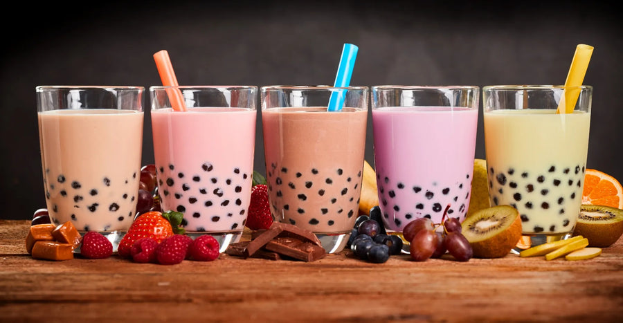 Drei Gläser Bubble Tea in rosa, braun und lila mit bunten Trinkhalmen und schwarzen Tapiokaperlen, umgeben von frischen Früchten und Gewürzen