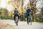 Zwei Radfahrer mit Helmen fahren lachend auf einem herbstlichen, umgeben von bunter Natur