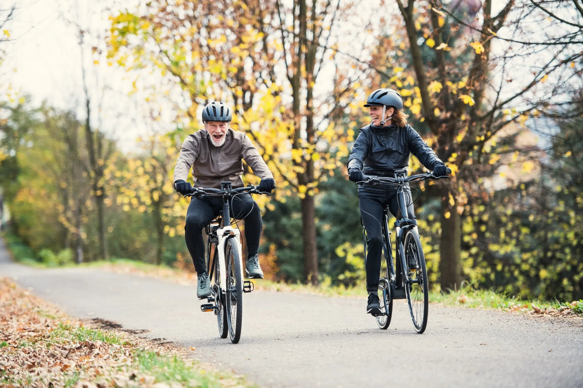 Zwei Radfahrer mit Helmen fahren lachend auf einem herbstlichen, umgeben von bunter Natur