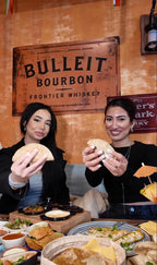 Zwei Frauen halten Tacos in die Kamera im Tequilaria Bochum vor rustikaler Wand mit Bulleit-Bourbon-Schild