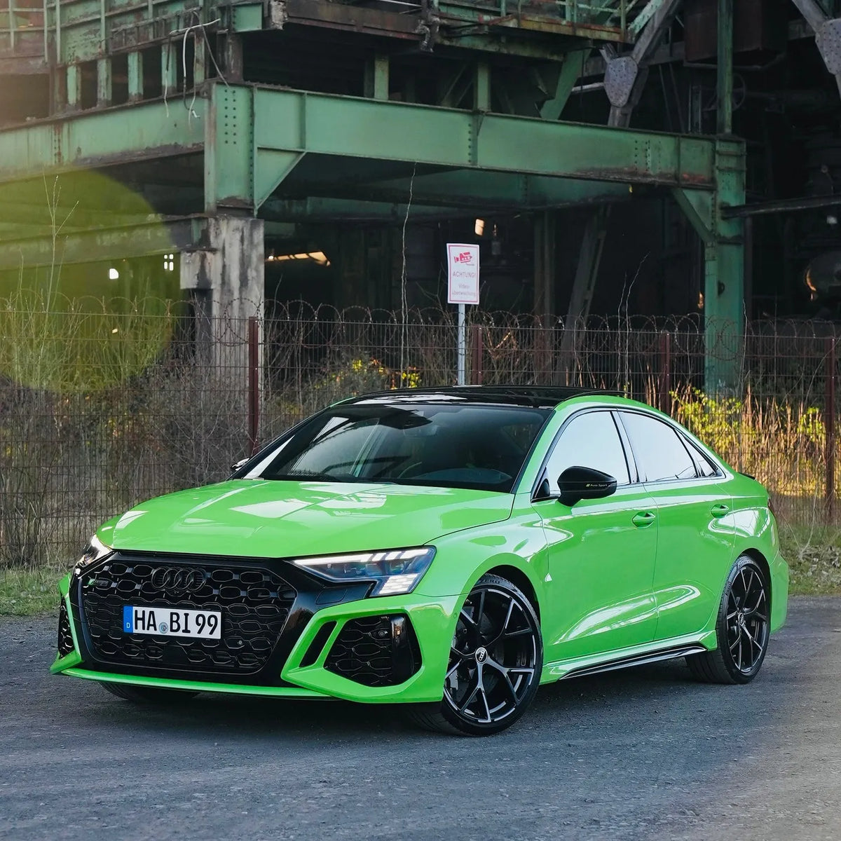 Audi RS3 Limousine in Neongrün, frontal-seitliche Ansicht vor industrieller Kulisse mit Sonnenreflexion