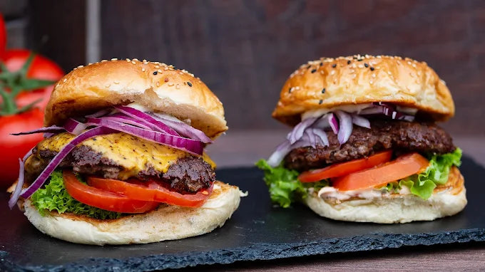 Zwei saftige Burger mit Cheddar, Tomate, Salat und roten Zwiebeln auf schwarzem Schiefer