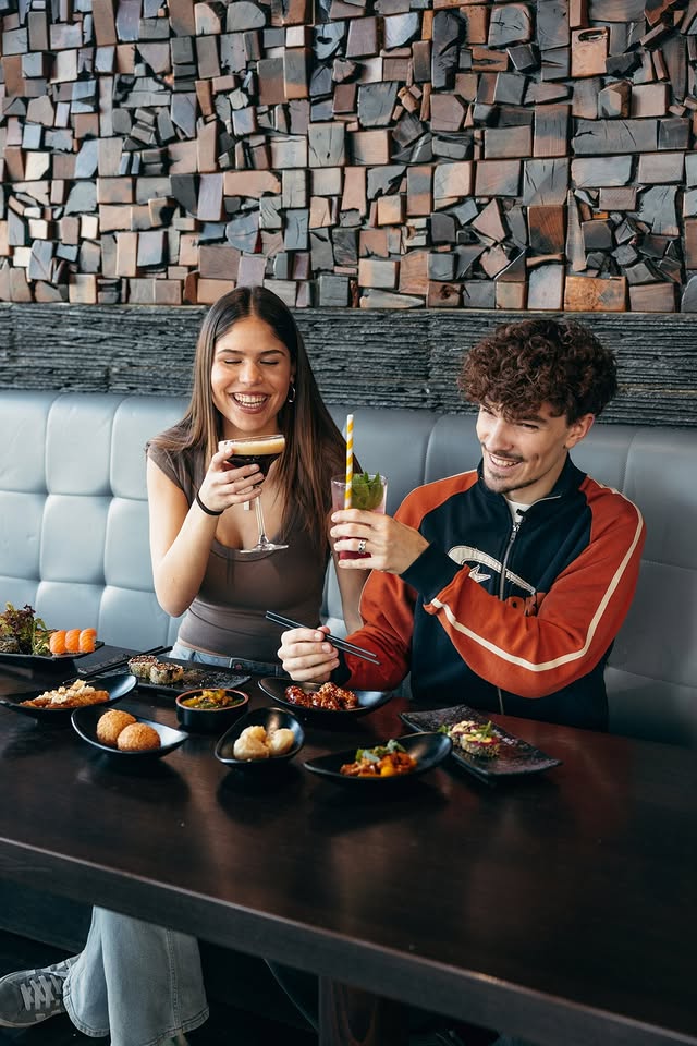 Junges Paar genießt verschiedene Tapas und Drinks in einem Restaurant mit moderner Holzwand-Deko. Auf dem Tisch stehen mehrere kleine Teller mit verschiedenen Gerichten und Getränken