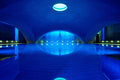 Weitwinkelaufnahme eines architektonisch markanten Hallenbads mit gewölbter Decke, intensiv blauer Beleuchtung, zentralem Oberlicht und stiller Wasseroberfläche mit gespiegeltem Lichtspiel.