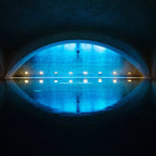 Frontale Ansicht eines ruhigen Hallenbeckens unter einem Gewölbe, mit blauer und gelber Akzentbeleuchtung, die sich auf der Wasseroberfläche spiegelt und eine meditative Stimmung erzeugt.