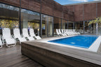 Modern gestalteter Außenbereich mit kleinem Pool auf blau gefliester Fläche, umgeben von weißen Liegestühlen und Holzdeck, vor einer Glasfassade mit farbigem Lichtspiel im Innenraum.