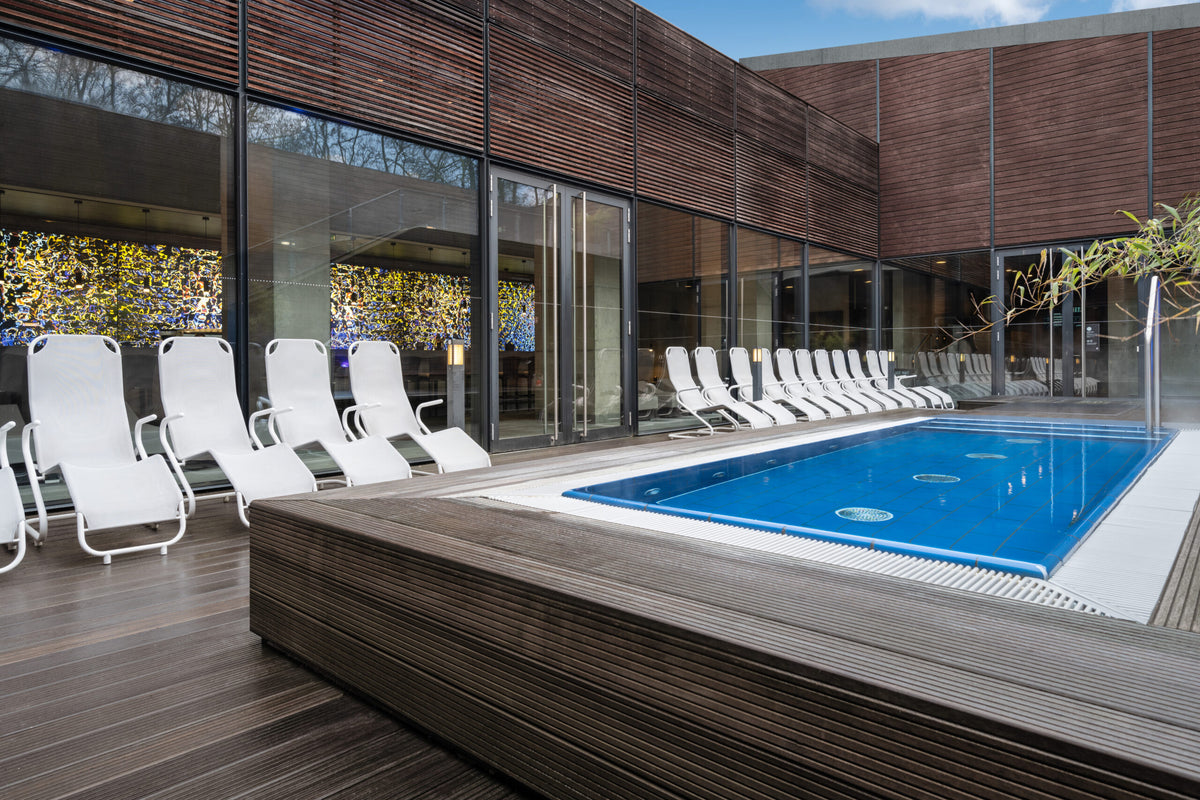 Modern gestalteter Außenbereich mit kleinem Pool auf blau gefliester Fläche, umgeben von weißen Liegestühlen und Holzdeck, vor einer Glasfassade mit farbigem Lichtspiel im Innenraum.