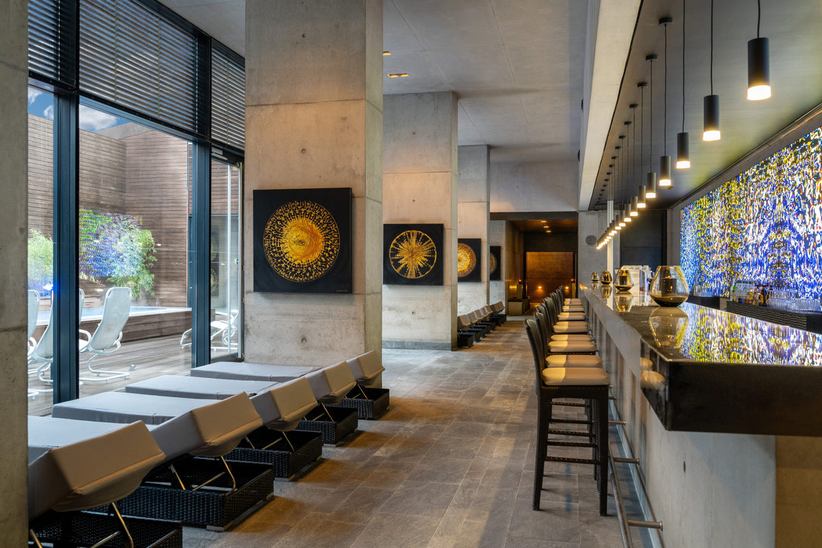 Eleganter Spa-Loungebereich mit Liegen am Fenster, modernen Kunstwerken an Betonwänden und stilvoll beleuchteter Bar mit leuchtendem Mosaik hinter dem Tresen.